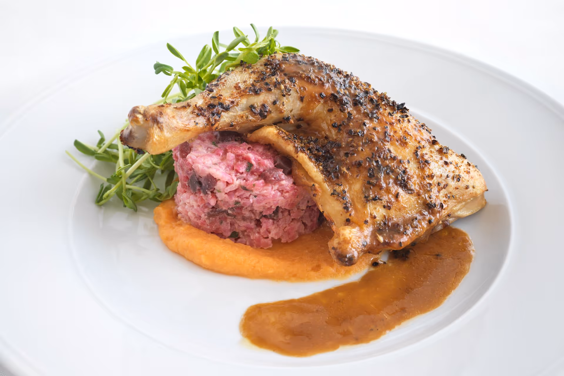 A plated gourmet dish featuring a seasoned and roasted chicken leg placed on top of a mound of pink rice or grain, accompanied by a dollop of orange sauce and garnished with fresh green herbs on a white plate.