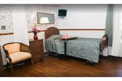 A single hospital-style bed with green bedding in a healthcare facility room. Next to the bed is a small overbed table with a pink box on it. There is a wooden dresser with a vase of flowers and a mirror above it. A cushioned armchair is positioned near the dresser. The room has wood flooring, floral wallpaper, and a window with green curtains.