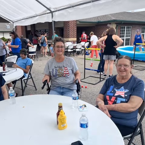 Two women sitting at a round white table outdoors under a canopy. One woman is wearing a gray shirt with USA printed on it and the other is wearing a blue shirt with LOVE and an American flag design. There are bottles of water, ketchup, and mustard on the table. In the background, other people are standing and sitting near a brick building, with some playing a lawn game and a small inflatable pool visible.