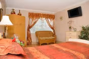 Warmly decorated bedroom with a red-orange bedspread, window with gold curtains, a wicker bench, dresser with plants, wall-mounted TV, and lamp.