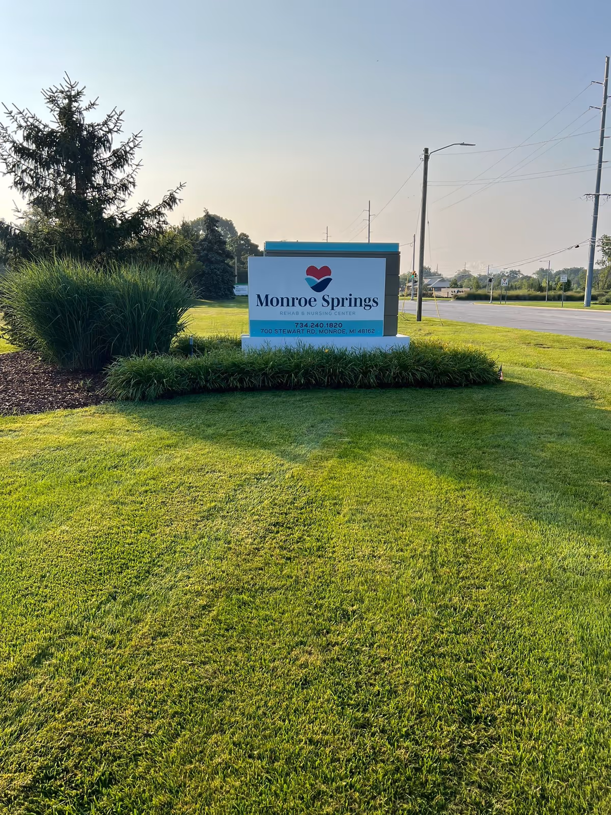 A large sign for Monroe Springs Rehab & Nursing Center situated on a well-maintained grassy area with bushes and trees nearby. The sign includes the facility's name, phone number, and address. A road and utility poles are visible in the background under a clear sky.