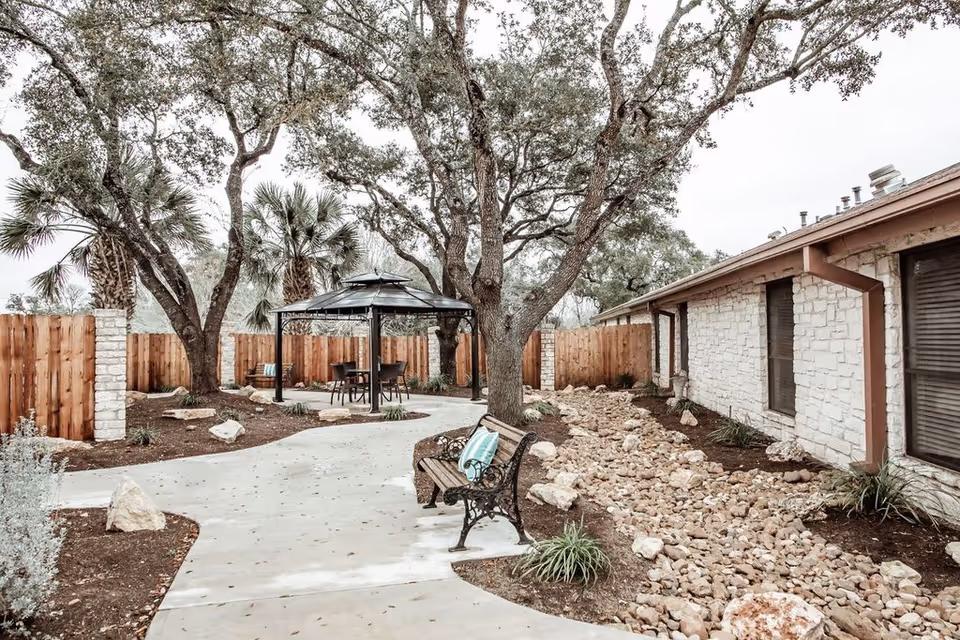 Outdoor garden area at Monument Hill Nursing and Rehabilitation Center featuring a paved walkway, a wooden bench with a cushion, large trees, a gazebo with chairs and a table, a stone-lined dry creek bed, and a wooden fence surrounding the space.