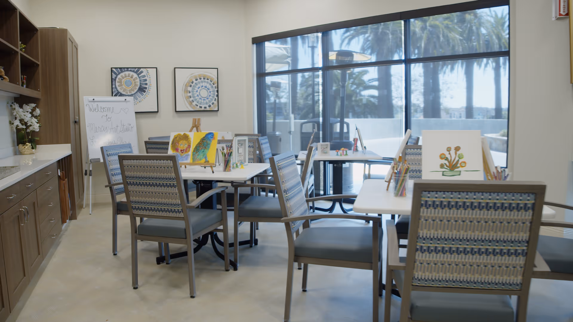 Bright interior activity room with tables, chairs, art supplies and paintings arranged near large windows.