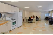 A spacious room with a kitchen area on the left featuring white cabinets, a stove, and a refrigerator. The room has a tiled floor with a checkered pattern in beige and light brown. In the background, there are several desks with chairs and office supplies, suggesting a multi-purpose space.