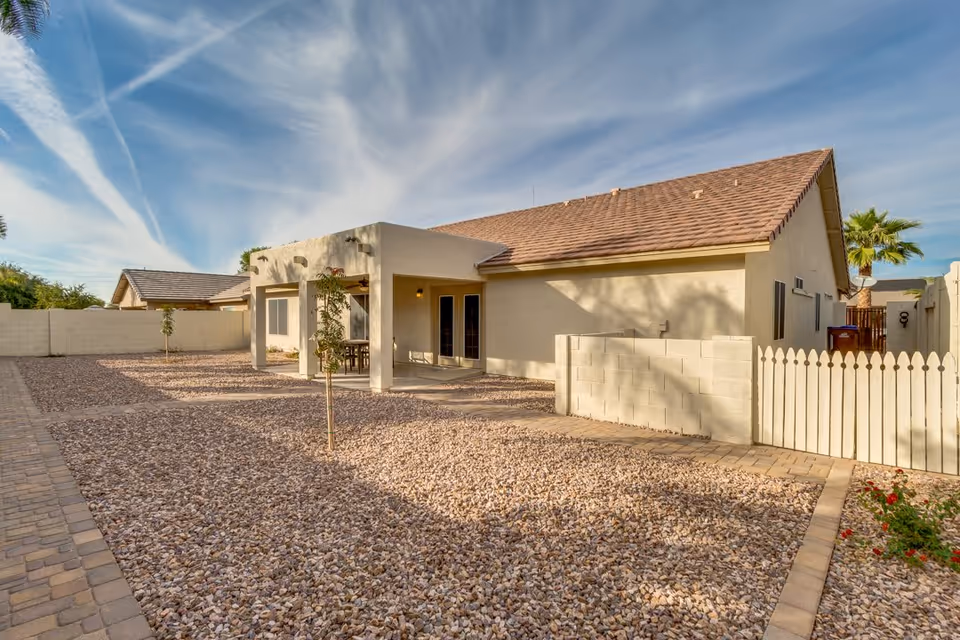 Exterior view of a single-story building with a covered patio area, surrounded by a yard covered in small rocks and a paved walkway. The building has a beige exterior with a brown shingled roof, a white picket fence on the right side, and a clear blue sky with wispy clouds overhead.