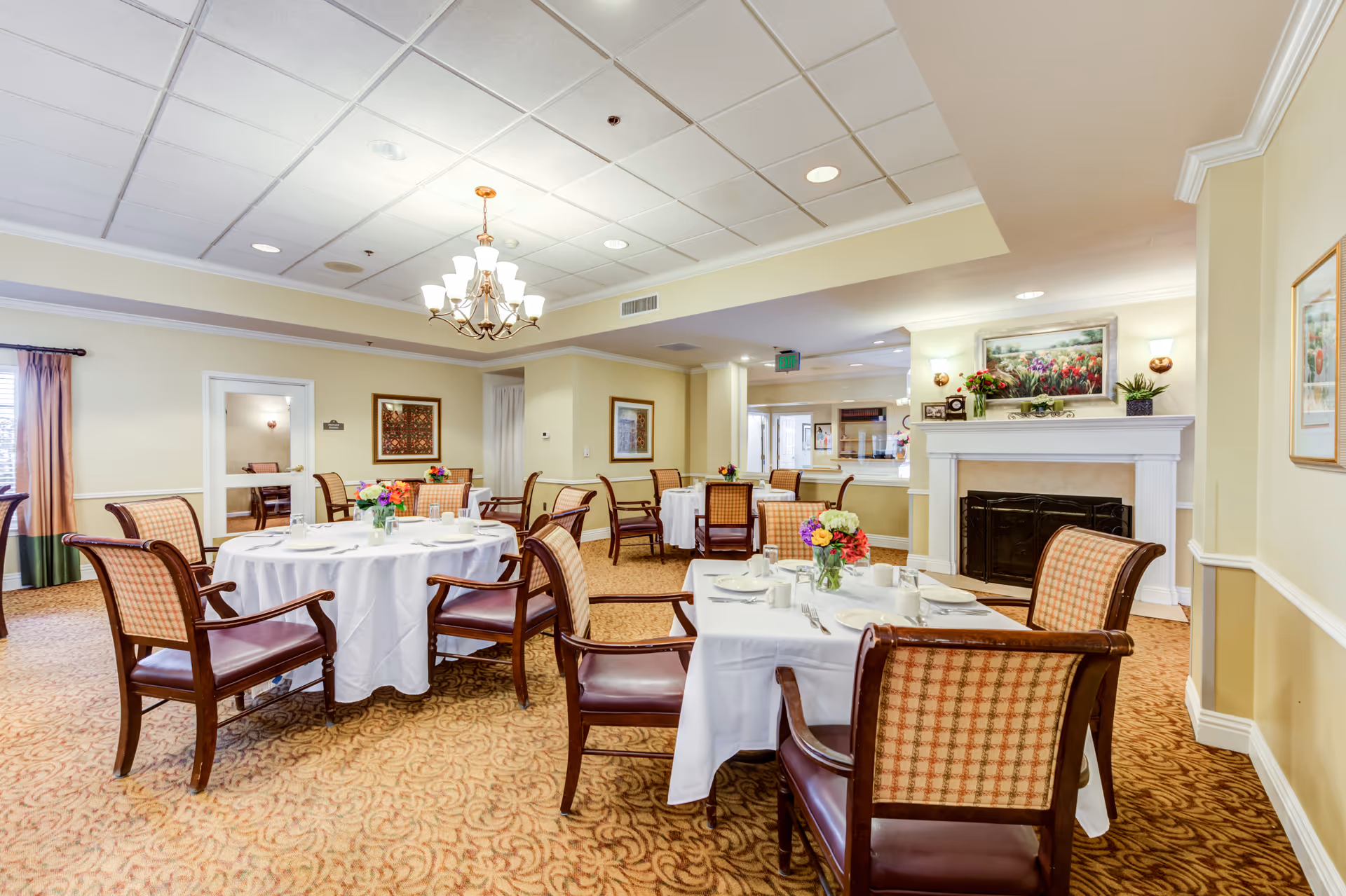 A bright and elegant dining room in a senior living facility with round tables covered in white tablecloths, set with plates, cups, and silverware. Each table has a small floral centerpiece. The room features patterned carpet, cushioned wooden chairs, a chandelier, framed artwork on the walls, and a fireplace with decorative plants and a painting above it.