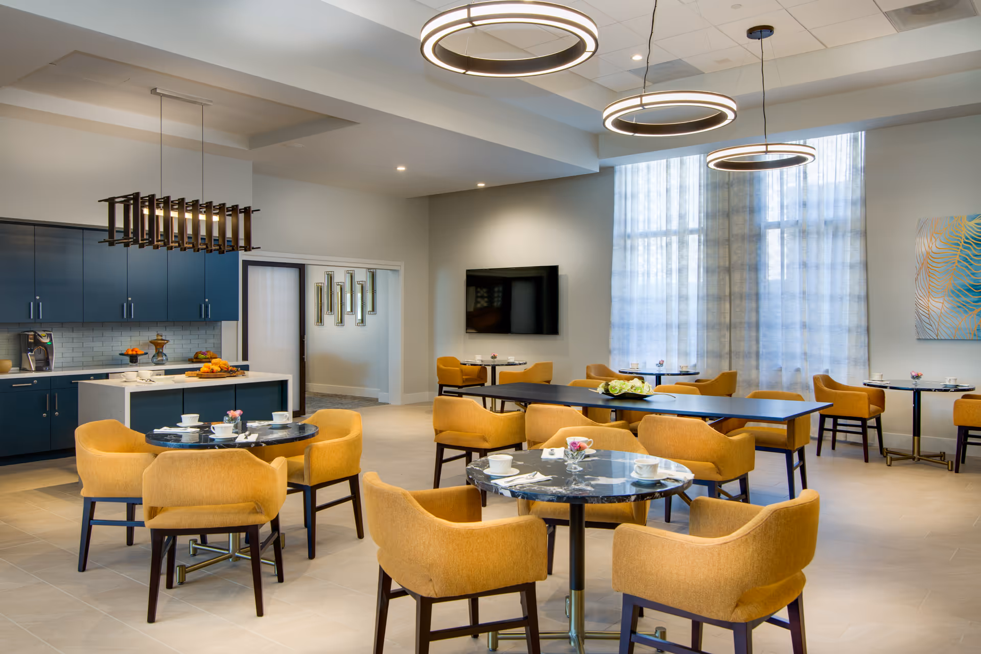 A modern dining area in a senior living facility with round black marble tables surrounded by mustard yellow upholstered chairs. The room features large windows with sheer curtains, contemporary circular pendant lights, a wall-mounted flat-screen TV, and a kitchen area with blue cabinets and a white countertop. The space is bright and inviting with neutral-colored flooring and walls.