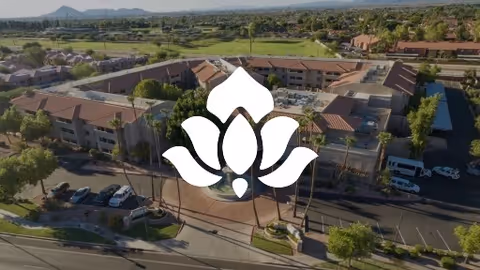Aerial view of a senior living facility with multiple buildings, parking lots, and surrounding greenery under a clear sky with mountains in the background.