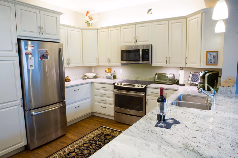 Modern kitchen with white cabinetry, stainless steel refrigerator, oven, microwave, and toaster. Granite countertops with a sink and a bottle of red wine with two wine glasses on a black napkin. Hardwood floor with a decorative rug in front of the cabinets.