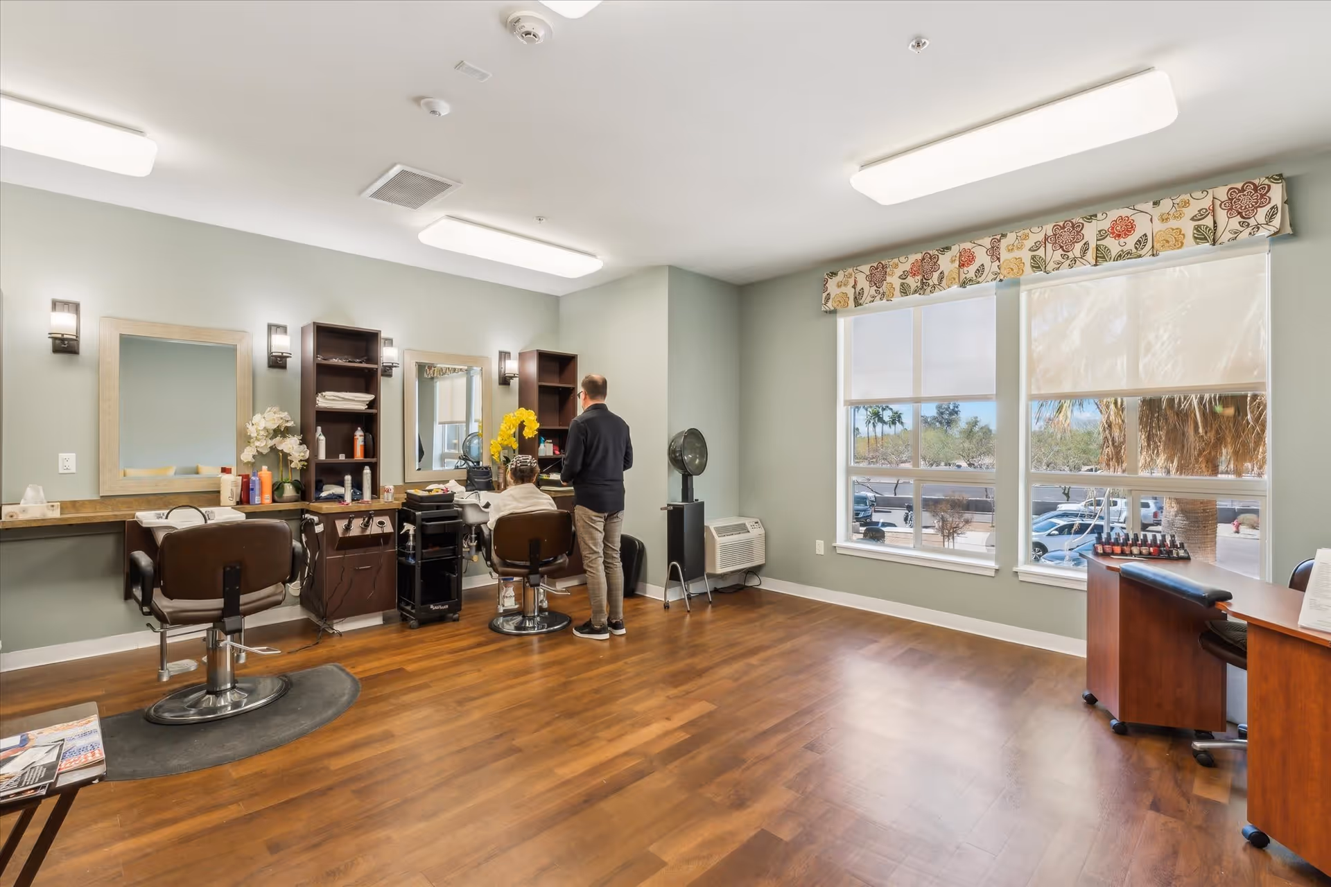 A spacious room with wooden flooring and light green walls, featuring two salon chairs in front of mirrors and shelves with hair care products. A person is standing behind another person seated in one of the chairs, possibly providing a hair service. Large windows with floral valances let in natural light, and a small table with nail polish bottles is visible near the windows.