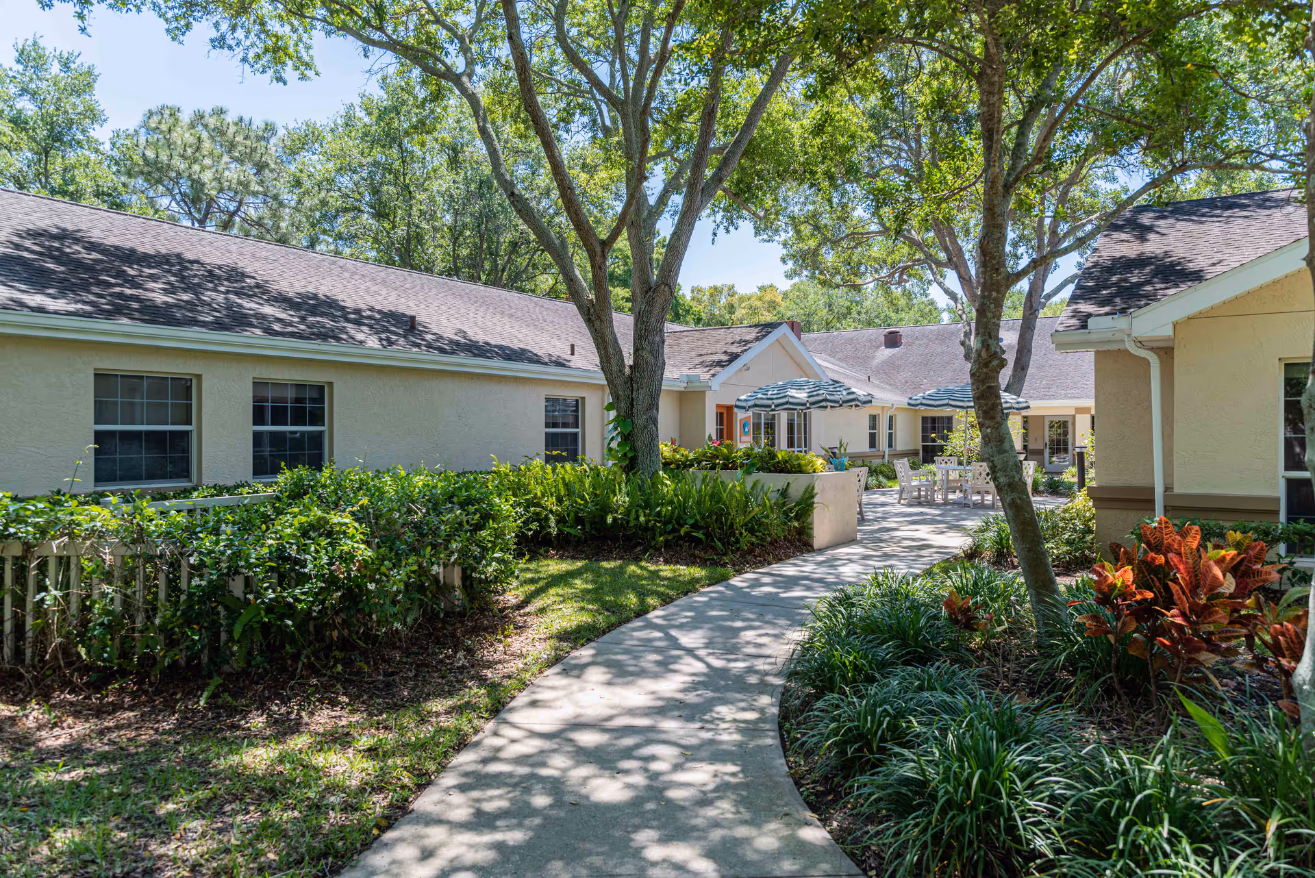 A sunny outdoor courtyard area at Arden Courts - ProMedica Memory Care Community (Largo) featuring a winding concrete pathway surrounded by green shrubs, trees, and plants. Beige buildings with multiple windows border the courtyard, and there are patio tables with umbrellas and chairs for seating.