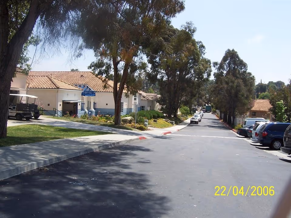 A paved road lined with parked cars on the right side and trees on both sides. Single-story buildings with tiled roofs and a golf cart are visible on the left side. The scene is sunny with clear skies.