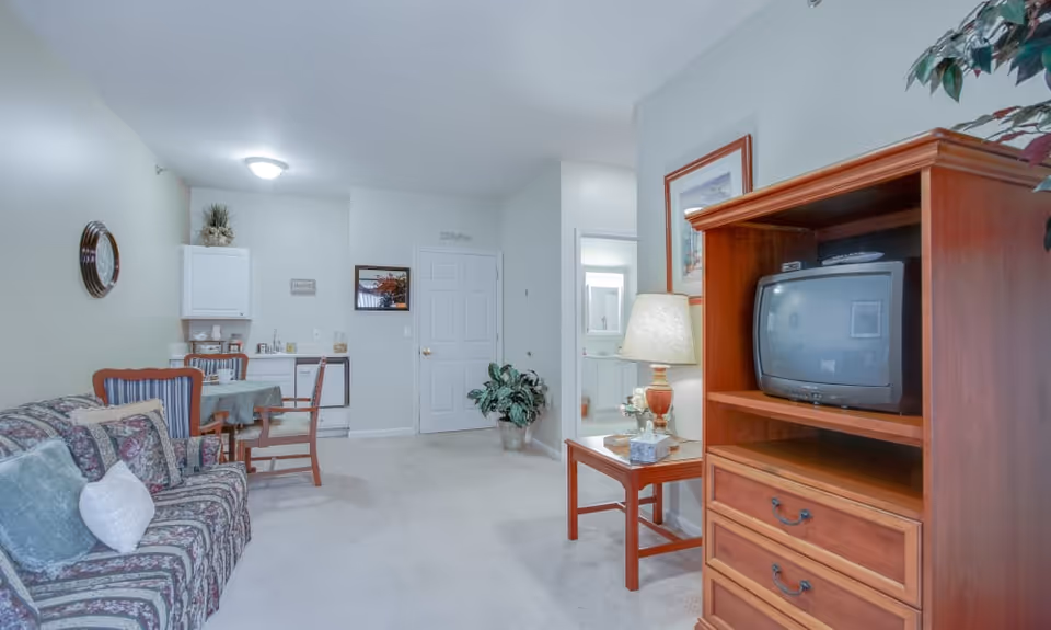 Interior view of a senior living facility room featuring a floral patterned sofa with cushions, a small dining table with chairs, a kitchenette area with cabinets and a sink, a wooden TV stand with an older style television, a side table with a lamp, and a potted plant near a closed door.