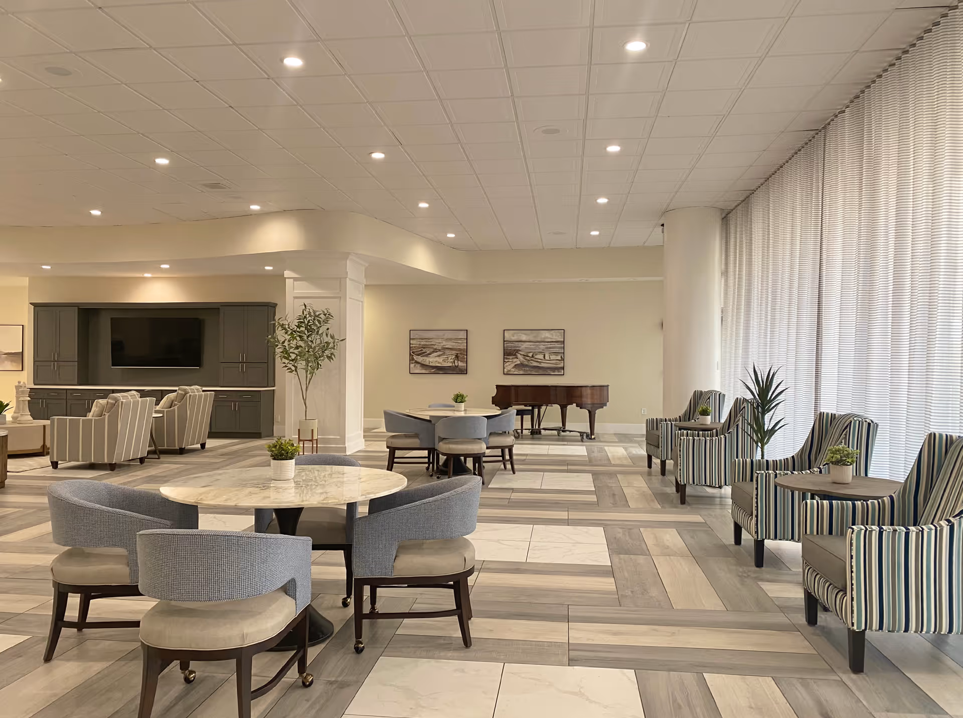 A spacious and well-lit senior living common area with round tables and upholstered chairs, striped armchairs along a large window with sheer curtains, a grand piano in the background, and a built-in entertainment center with a television. The flooring features a mix of wood and tile patterns.