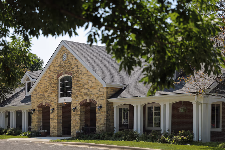 Exterior view of a single-story assisted living facility building with a stone facade and a gabled roof, partially framed by green tree branches.