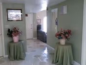 Interior view of a hallway in a senior living facility with two small round tables covered with green cloths, each holding a pink flower pot with artificial flowers. The floor is tiled with a light pattern, and there is a framed picture on the left wall. At the end of the hallway, there is a seating area with a striped couch near a window letting in natural light.