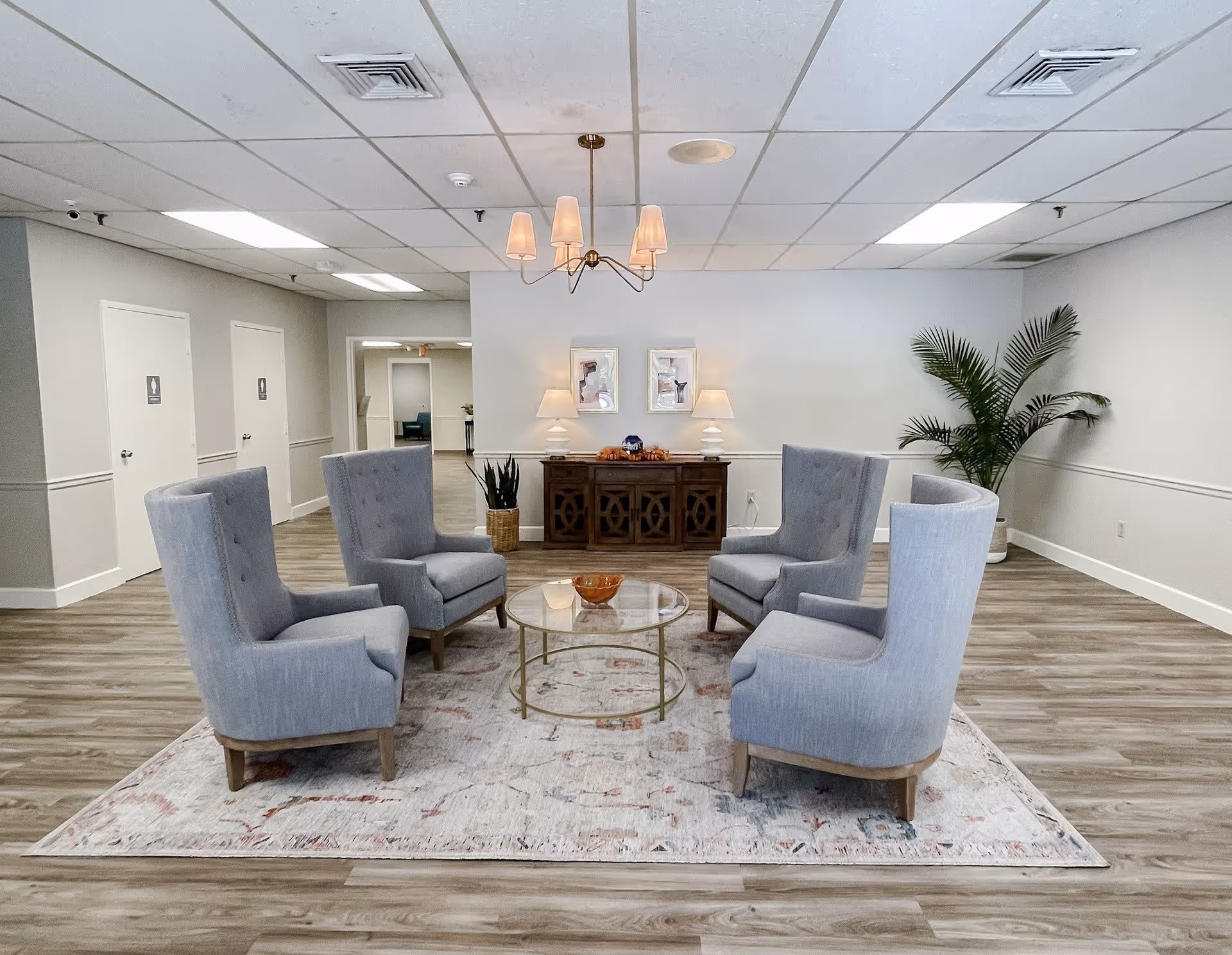 A senior living facility common area with four gray upholstered armchairs arranged around a round glass coffee table on a patterned rug. The room has light wood flooring, white walls, and a white ceiling with recessed lighting and a chandelier. A wooden sideboard with two table lamps and framed artwork is against the back wall, with two potted plants on either side.
