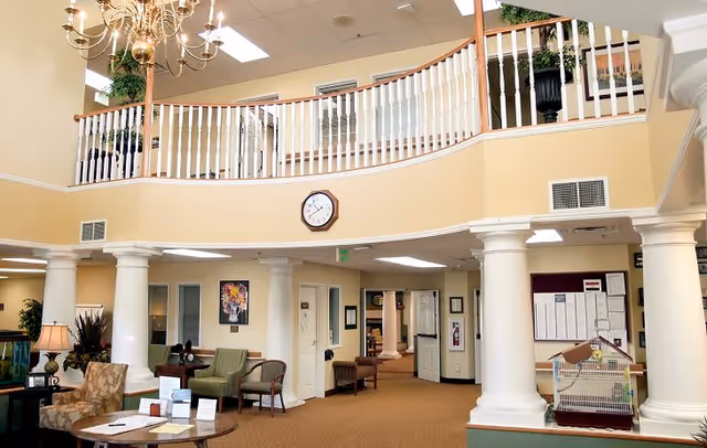 Interior view of a senior living facility lobby with beige walls, white columns, a chandelier, a clock on the wall, seating area with chairs and tables, and a birdcage on a stand. There is a second-floor balcony with white railings and plants.