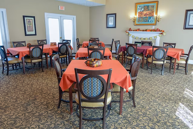 Dining room with several square tables covered in red tablecloths, wooden chairs, patterned carpet, and a decorated fireplace with framed artwork.