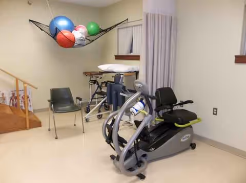 Rehabilitation room with stationary bike, exercise balls, chairs, and medical equipment.