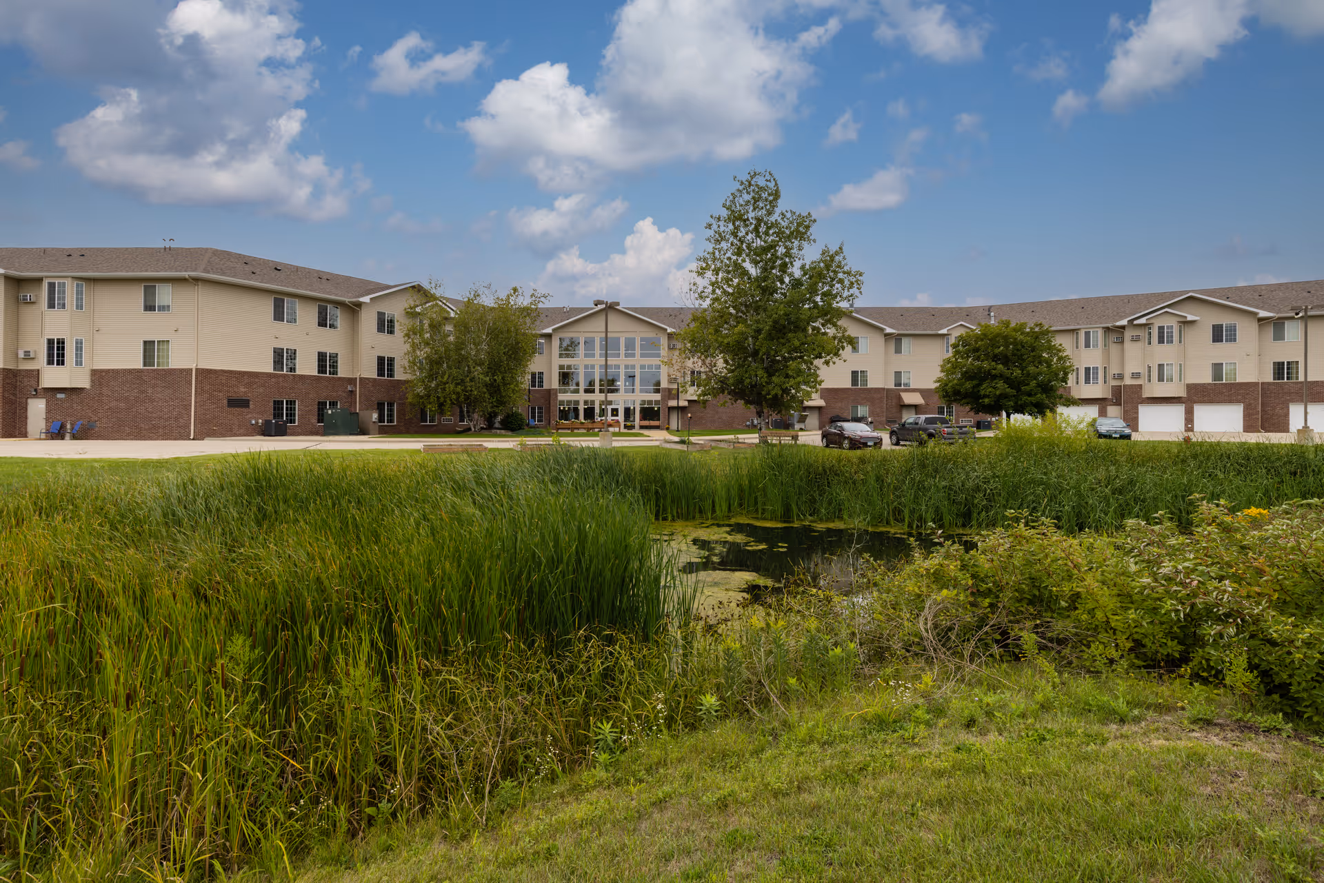 Exterior view of Prairie Meadows Senior Living building with a pond and tall grass in the foreground, trees near the building, and a partly cloudy sky above.