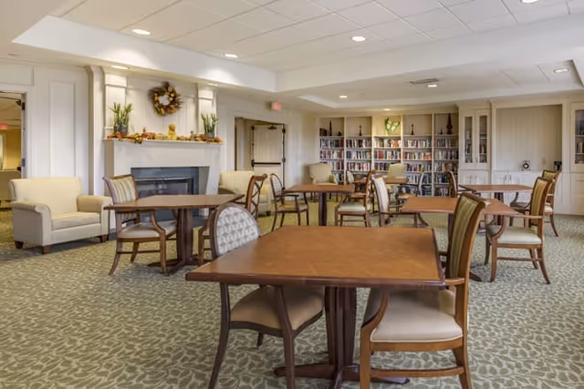 Spacious common room with multiple wooden tables and chairs, a fireplace, armchairs, and bookshelves.