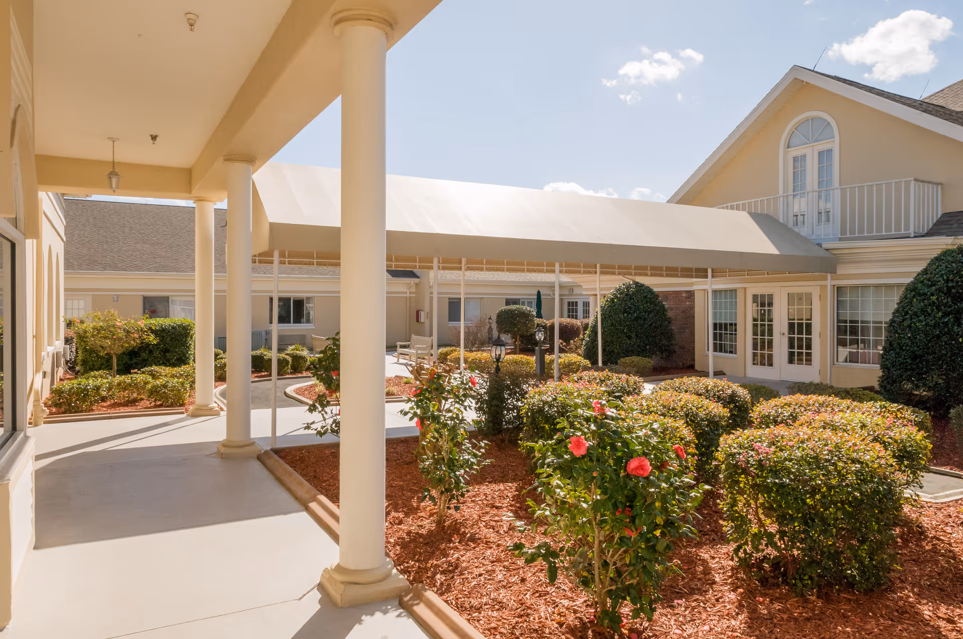 Outdoor courtyard area of a senior living facility with a covered walkway supported by white columns, neatly trimmed bushes, flowering plants, and a building with large windows and a balcony under a clear blue sky.