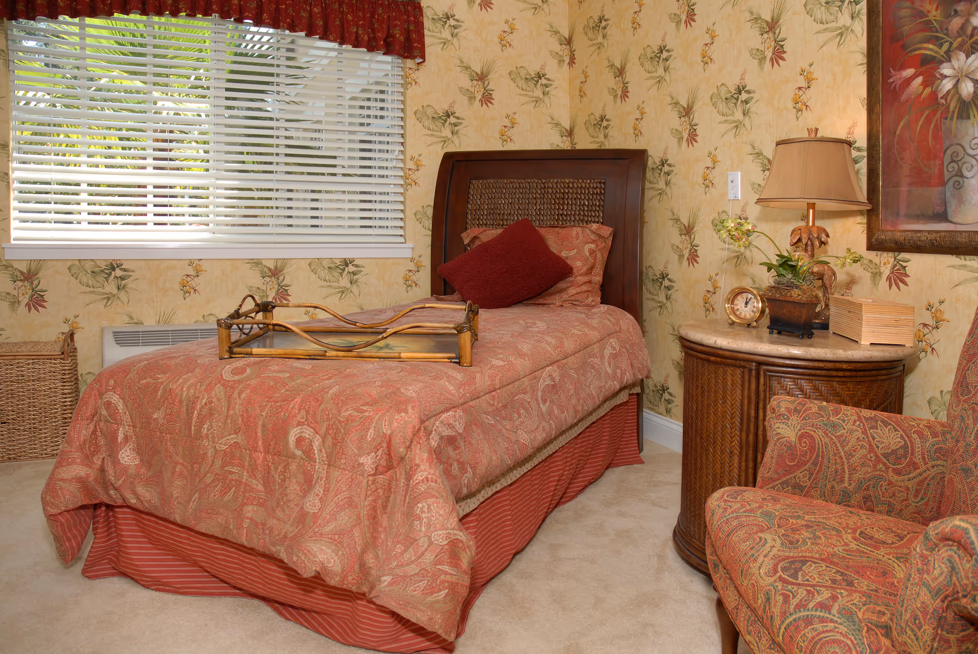 A cozy bedroom with a single bed covered in a red patterned bedspread and a matching pillow. There is a wooden tray on the bed. The room has floral wallpaper, a window with white blinds, a wicker nightstand with a lamp, clock, and plant, and a patterned armchair in the corner.