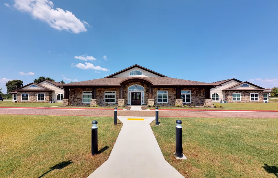 Front exterior view of a single-story building with a stone facade and a brown roof under a clear blue sky. A concrete walkway leads to the main entrance, flanked by four black lamp posts. The building has multiple windows and a central arched doorway.