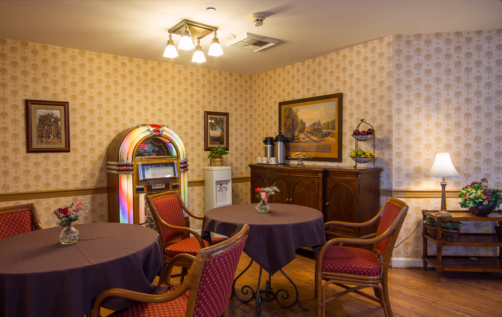 A cozy interior room with two round tables covered with dark tablecloths, surrounded by red cushioned wooden chairs. The room features vintage wallpaper, a colorful jukebox, framed artwork, a wooden sideboard with coffee dispensers and cups, a water cooler, and a small table with a lamp and flower arrangements.