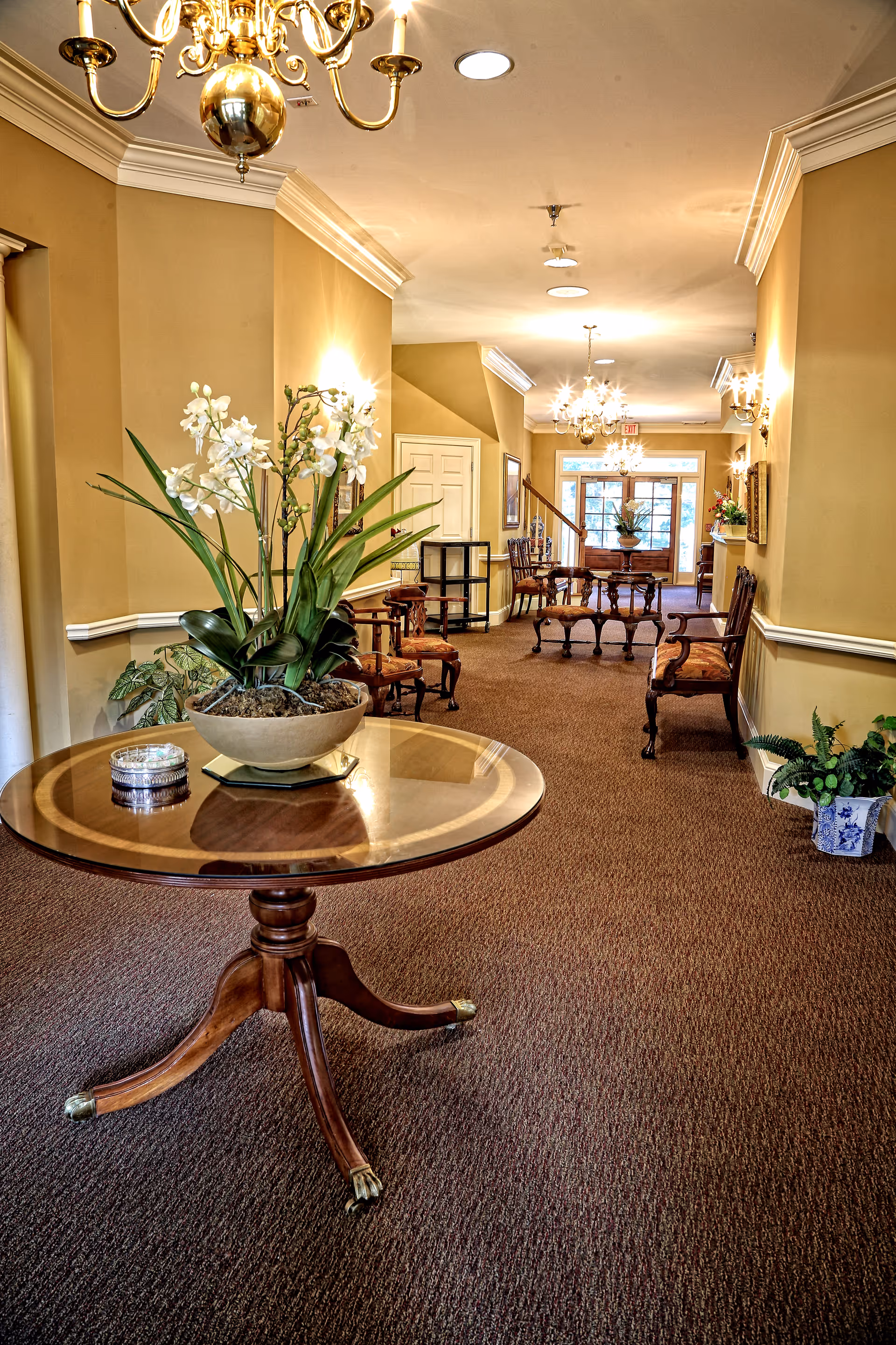 A warmly lit hallway in a senior care facility with beige walls and carpeted floor. There is a round wooden table with a potted plant and a decorative item on it in the foreground. Along the walls are several wooden chairs with upholstered seats. Chandeliers and wall sconces provide lighting, and there is a staircase and glass doors visible at the end of the hallway.