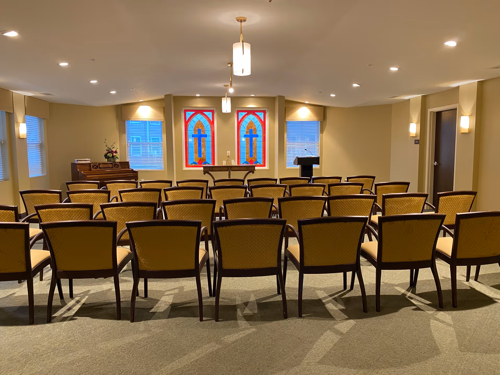 A room set up with rows of yellow cushioned chairs facing a small stage area with a podium and a table. Behind the stage are two stained glass windows with cross designs and two regular windows with blinds. A piano is positioned to the left side of the stage. The room has beige walls, carpeted floor, and ceiling lights.