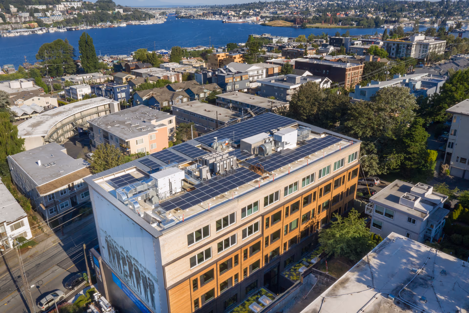 Aerial view of a multi-story building with solar panels installed on the rooftop, surrounded by other residential and commercial buildings near a large body of water with boats and greenery in the background.