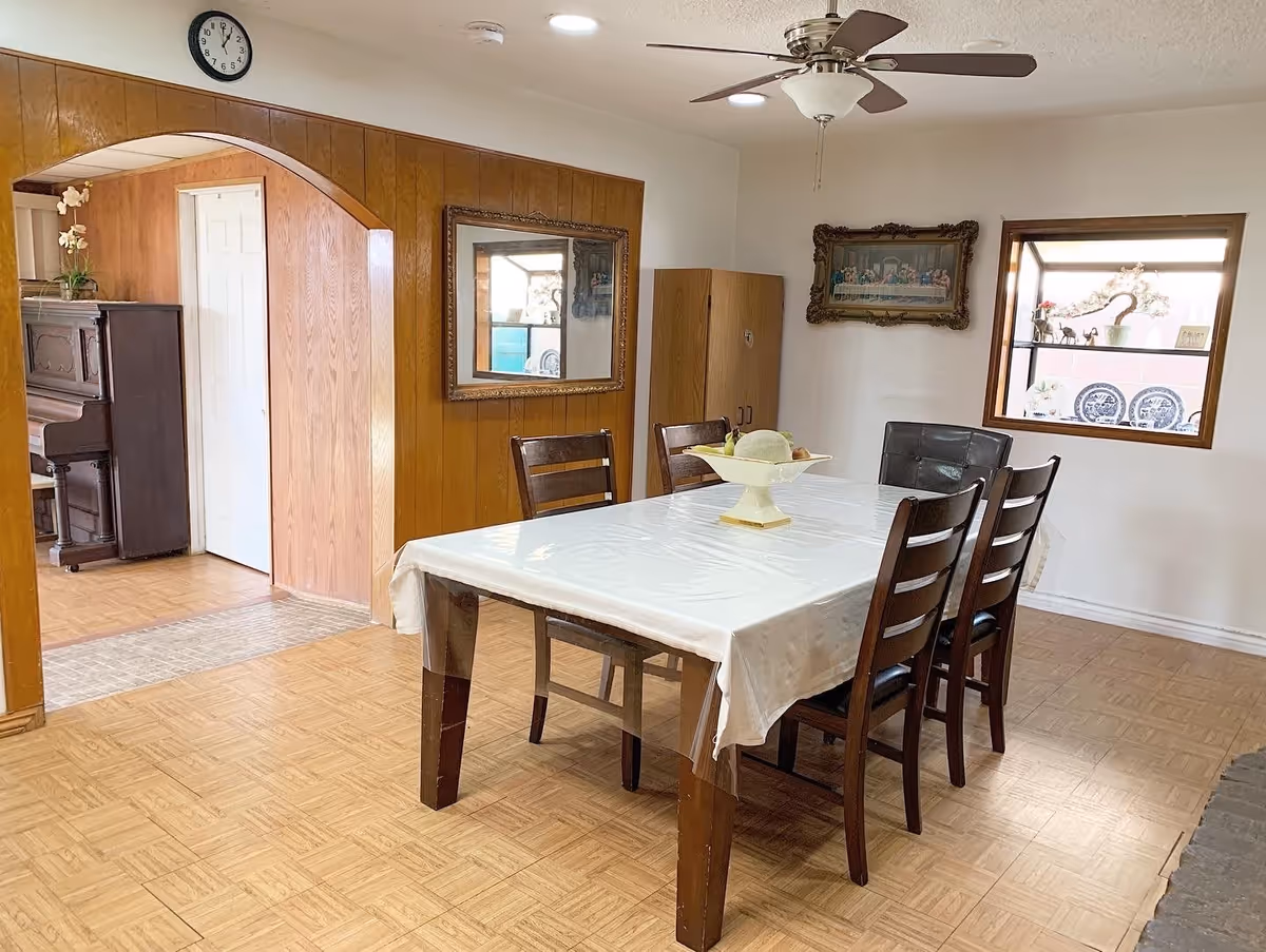 Dining room with a rectangular table covered with a white tablecloth and six wooden chairs. A ceiling fan with light is above the table. The room has wood-paneled walls on one side and white walls on the other. There is a large mirror on the wood-paneled wall and a framed picture on the white wall. A window with decorative plates and figurines on the sill is visible. An archway leads to another room with a piano and a closed white door. The floor has a light wood pattern.