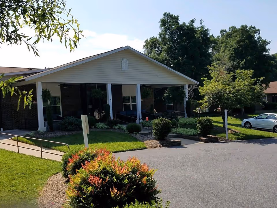 Exterior view of a single-story building with a covered entrance supported by white columns. There is a paved driveway and a sidewalk with a handrail leading to the entrance. The surrounding area has well-maintained green lawns, bushes, and trees. A white car is parked on the right side near the building.