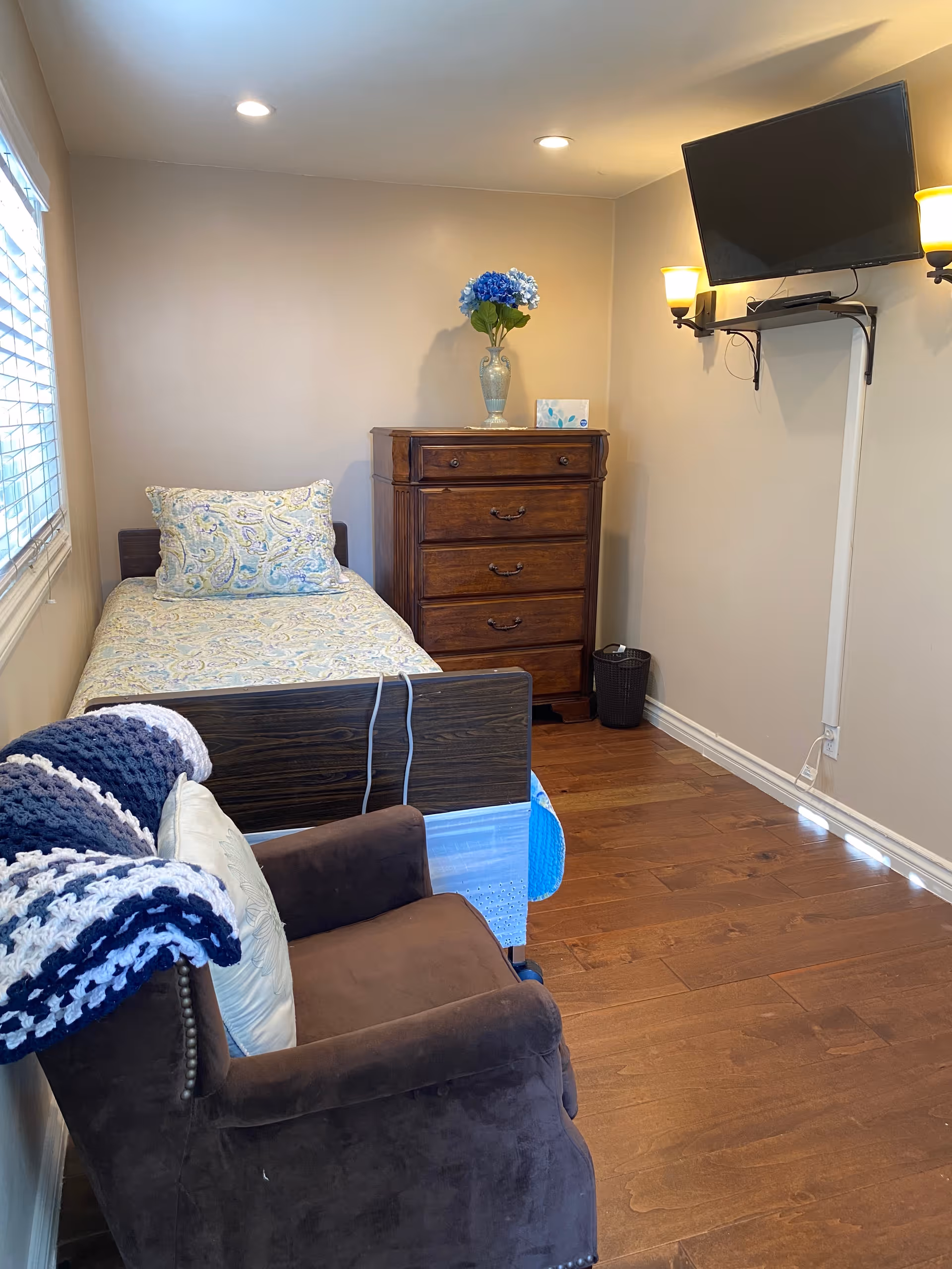 A small bedroom with a single bed covered in a patterned bedspread and pillow, a wooden chest of drawers with a vase of blue flowers and a box of tissues on top, a brown upholstered armchair with a crocheted blanket draped over it, a wall-mounted flat screen TV, and two wall sconces providing light. The room has wooden flooring and a window with blinds on the left side.