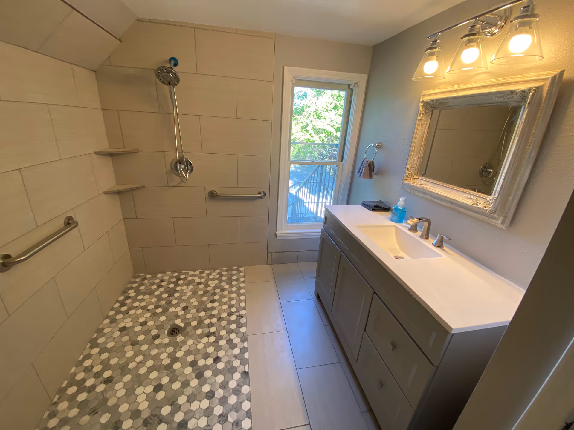 A modern bathroom featuring a walk-in shower with hexagonal tile flooring and beige wall tiles. The shower has a handheld showerhead and two built-in corner shelves. There are two grab bars installed on the shower walls. Next to the shower is a window letting in natural light. On the right side, there is a gray vanity with a white countertop, a sink, a faucet, a soap dispenser, and a decorative framed mirror above it. A three-light fixture is mounted above the mirror.