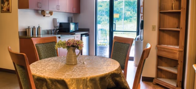 A cozy dining area in Oakwood Terrace featuring a round table covered with a patterned tablecloth and a vase of flowers in the center. Four wooden chairs with green upholstered backs surround the table. In the background, there is a small kitchenette with a microwave, sink, and cabinets. A glass door leads to an outdoor area with greenery visible outside. A wooden shelving unit is positioned to the right of the door.
