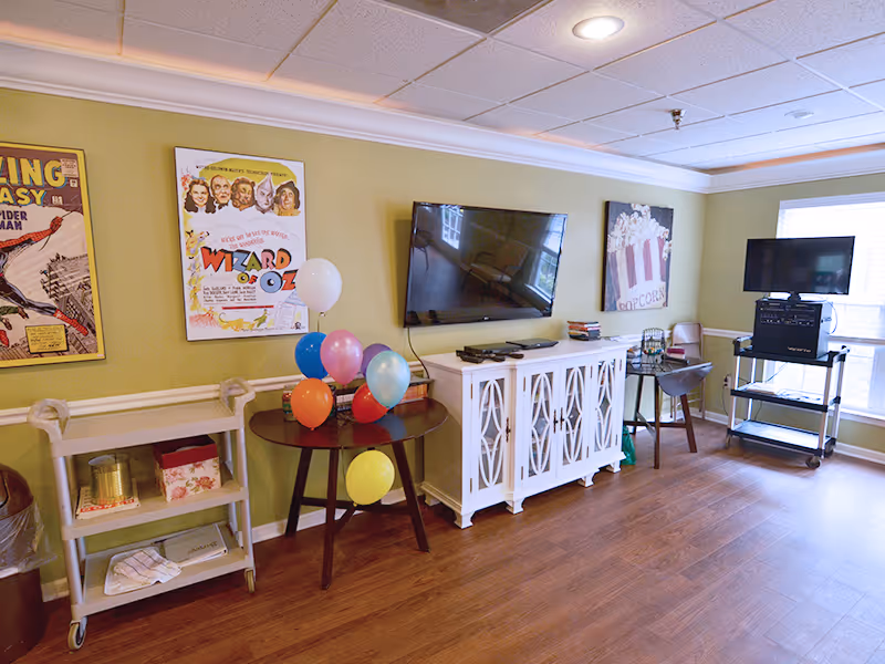 A cozy room with light green walls and wooden flooring featuring a wall-mounted flat screen TV above a white cabinet. There is a small round table with colorful balloons, a rolling cart with various items, and two framed posters on the wall, one of which is a Wizard of Oz movie poster. A TV on a stand is positioned near a window with blinds, and a chair is placed nearby.