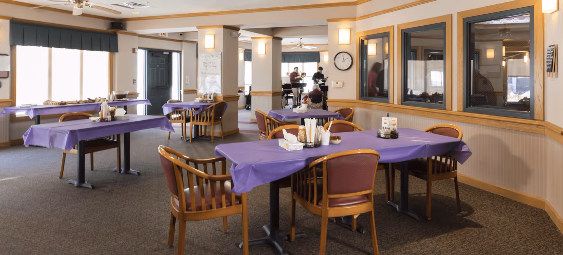 A dining area in an assisted living facility with tables covered in purple tablecloths and wooden chairs around them. In the background, there are large windows with green valances, a clock on the wall, and a small group of people playing musical instruments near the back of the room.