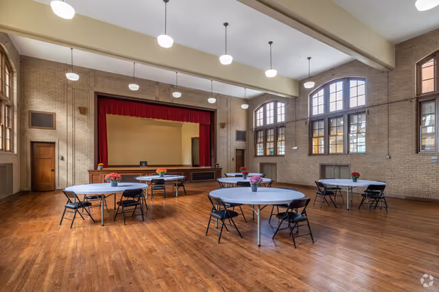 A large room with wooden floors and high ceilings featuring round tables covered with white tablecloths and black folding chairs. Each table has a small flower arrangement. At the far end of the room is a stage with a red curtain and a wooden podium. Large arched windows let in natural light.