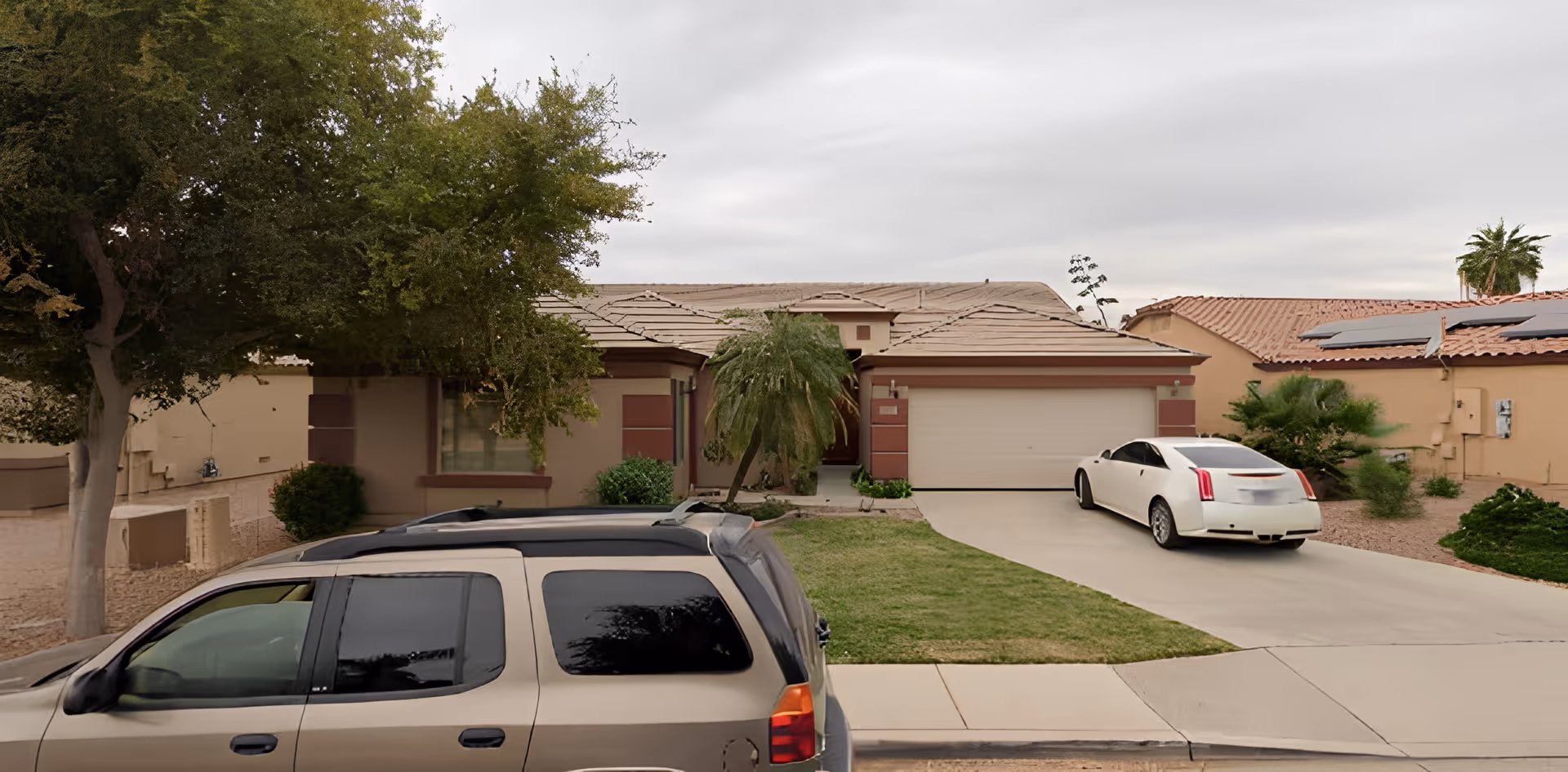 Front exterior view of a single-story residential house with a tiled roof, a two-car garage, a driveway with a white car parked on it, a green lawn, and trees on either side of the house.