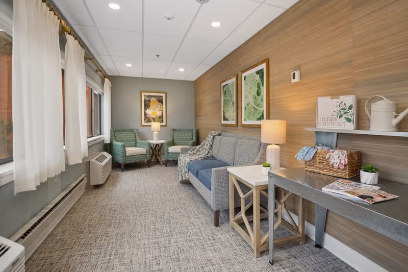 A cozy sitting area in a senior living facility with a gray sofa, two green patterned armchairs, a small round table with a lamp, and a long metal table with a lamp, a basket, and a book. The walls are decorated with framed artwork and a wooden panel accent wall. Large windows with white curtains let in natural light.