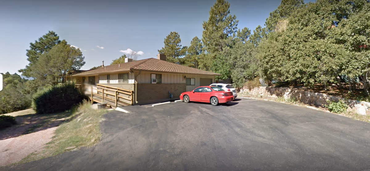 Exterior view of a single-story assisted living facility building with a brown roof and beige walls, surrounded by trees and greenery. There is a paved parking area in front with two parked cars, one red and one white. A wooden ramp leads to the entrance of the building.