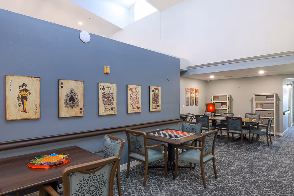 Bright common activity/dining room with tables and chairs, game boards on tables, card-themed wall art, and shelving.