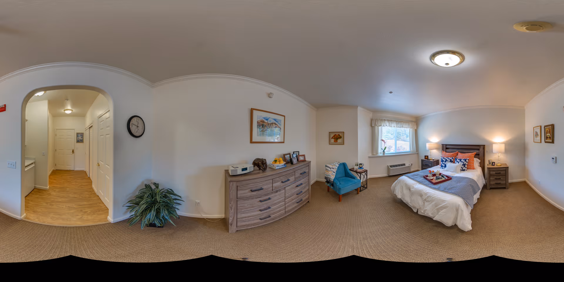 A cozy bedroom in a senior living facility featuring a neatly made bed with white and blue bedding, a wooden headboard, two bedside tables with lamps, a blue armchair with a side table, a dresser with decorative items and framed photos, a window with a valance letting in natural light, and a hallway leading to other rooms with wooden flooring.