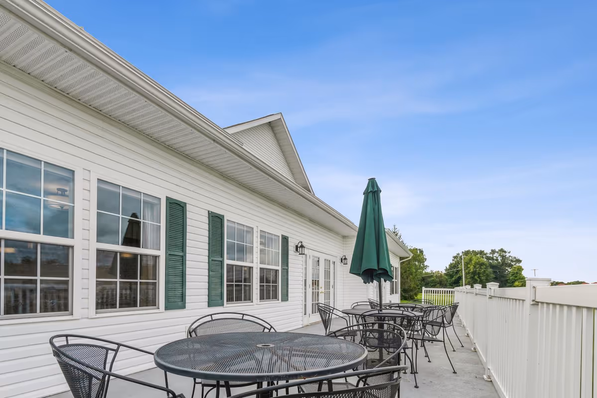 Outdoor patio area at Jefferson Manor with multiple round metal tables and chairs, some tables have closed green umbrellas. The patio is adjacent to a white building with green shutters and multiple windows under a clear blue sky.