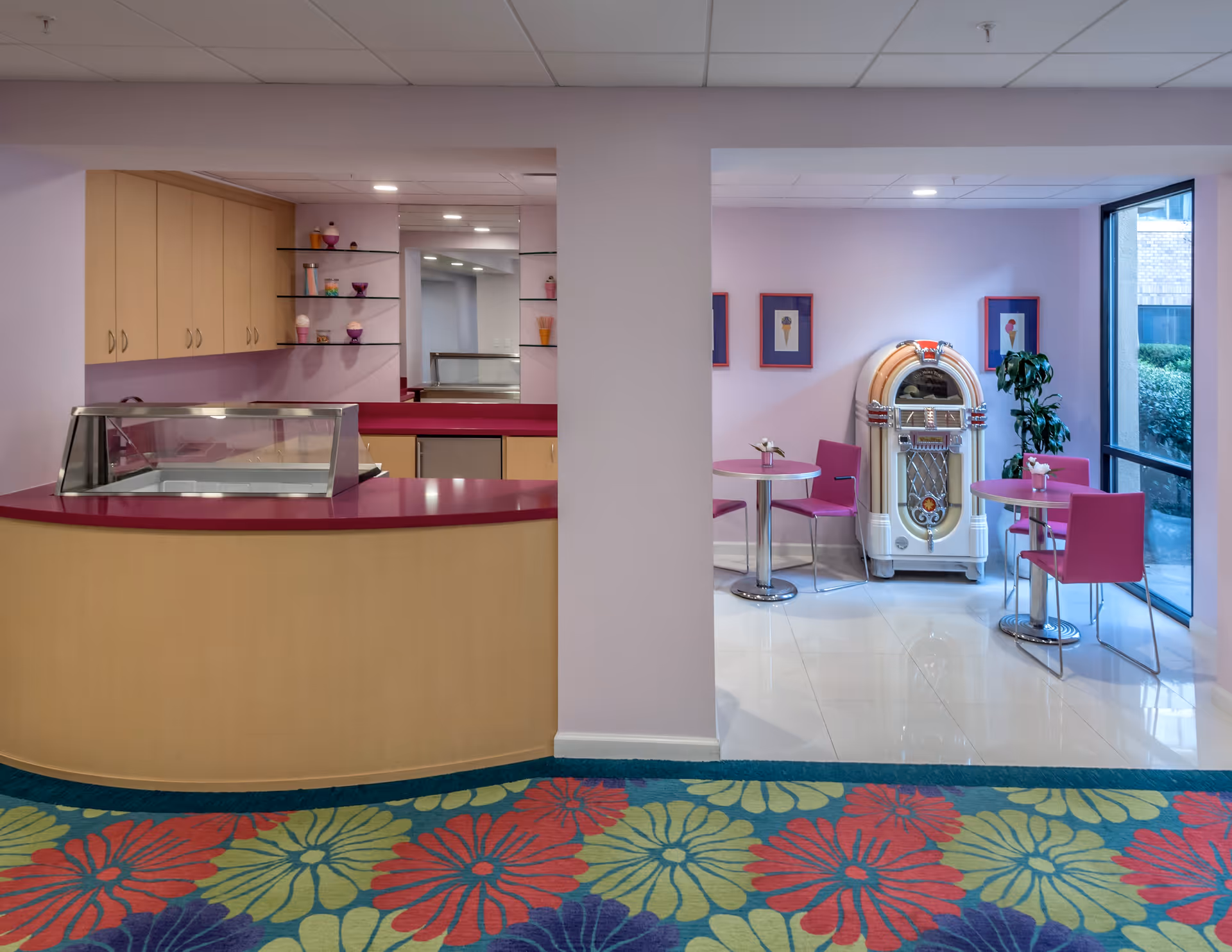 Interior view of a senior living facility area featuring a curved counter with a glass display case on the left and a small seating area on the right with pink chairs and round tables. A vintage jukebox stands against the wall, which is decorated with framed pictures of ice cream cones. Large windows provide natural light, and the floor has a colorful floral carpet in the foreground.
