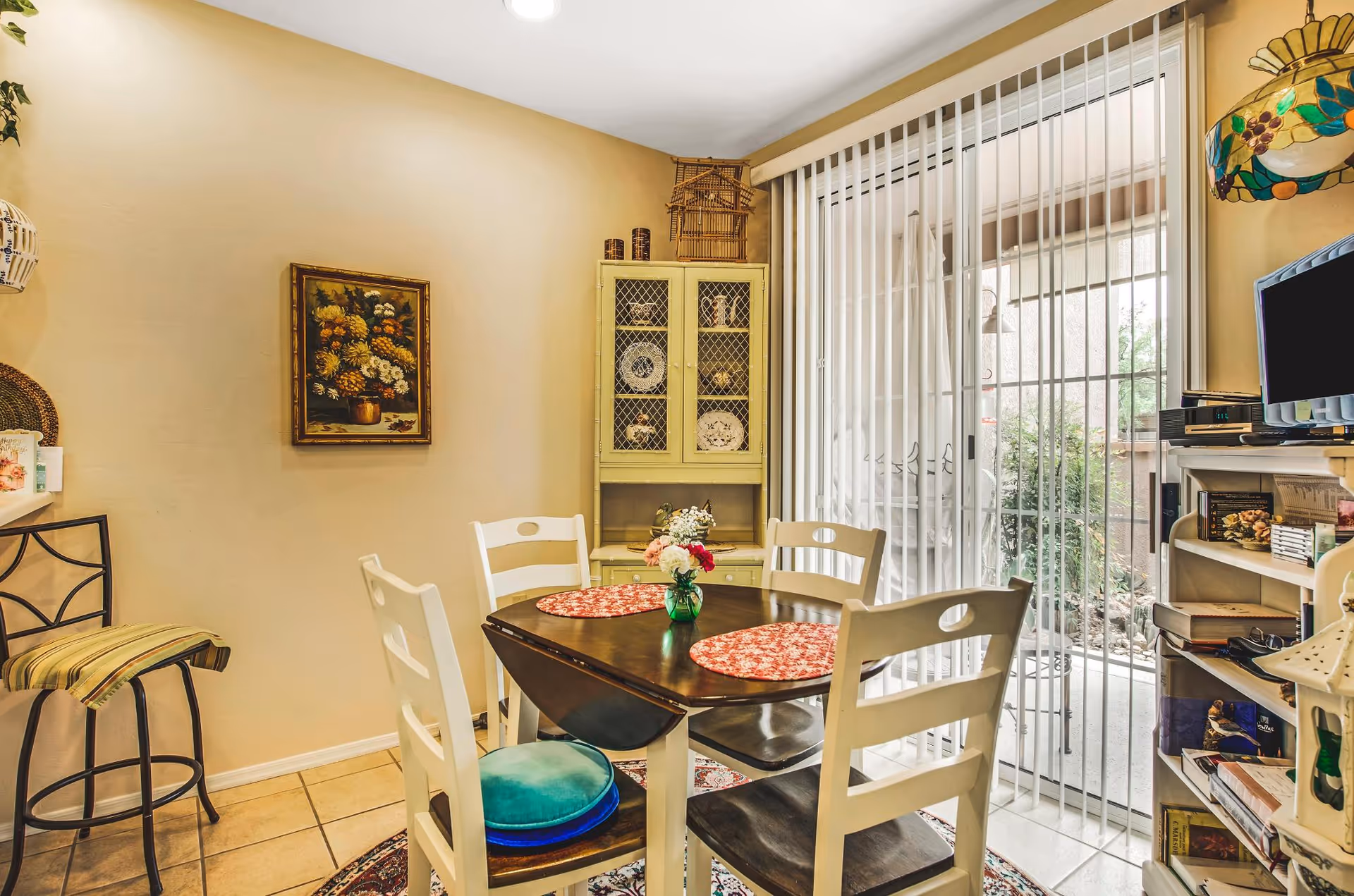 A cozy dining area with a dark wooden table and four white chairs, two with colorful cushions. The table has two red patterned placemats and a small vase with flowers. Behind the table is a yellow cabinet with decorative plates and items. To the right, vertical blinds cover a sliding glass door leading outside. On the right side, there is a white shelving unit with books and a small TV on top. The walls are light beige, and a framed floral painting hangs on the left wall.