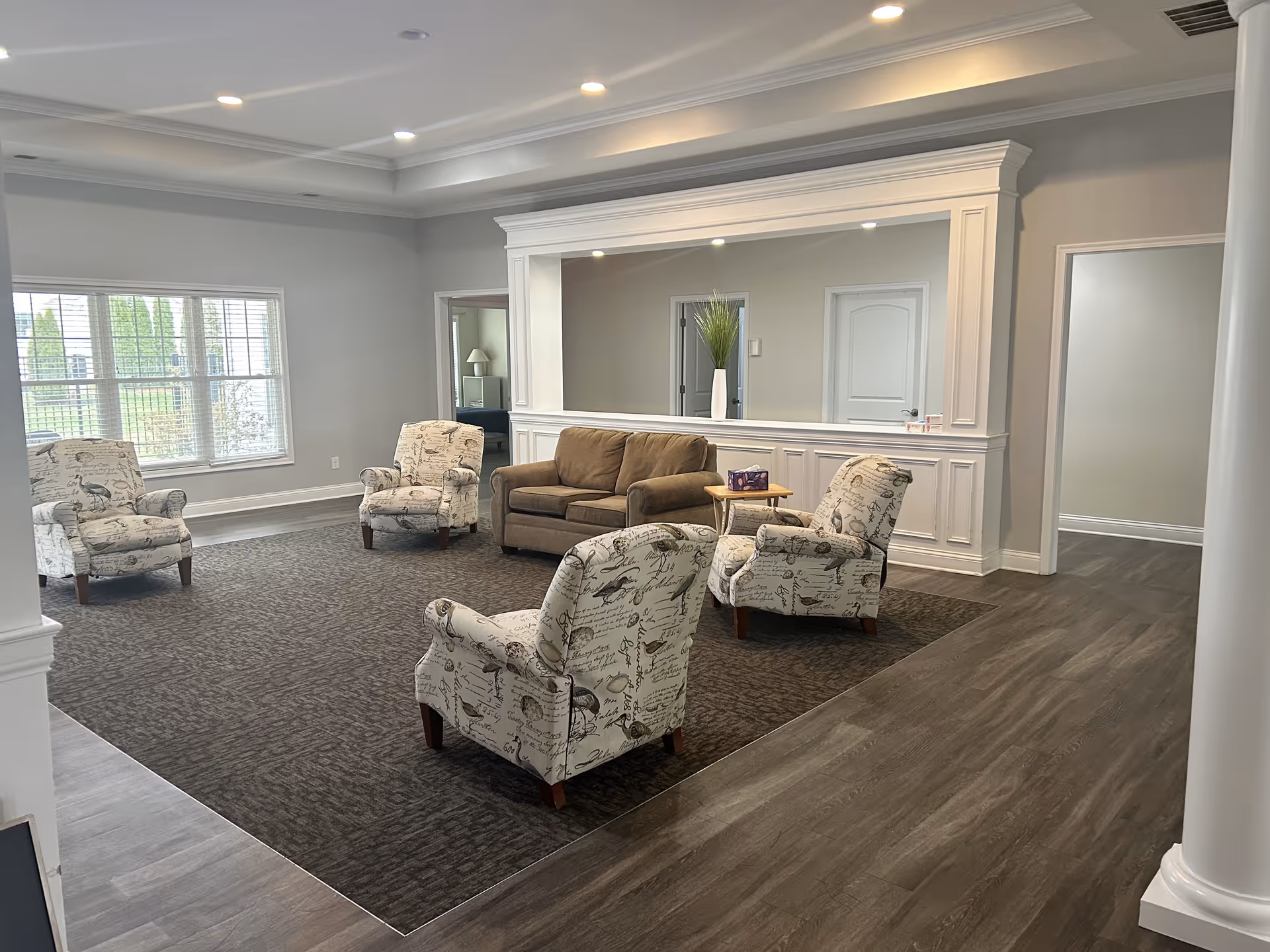 A spacious living room area with four patterned armchairs and a brown loveseat arranged on a dark carpet. The room has light gray walls, a large window with white blinds, and a large decorative mirror framed with white molding on one wall. There is a small wooden side table with a tissue box next to the loveseat. The floor outside the carpeted area is a wood-like laminate.